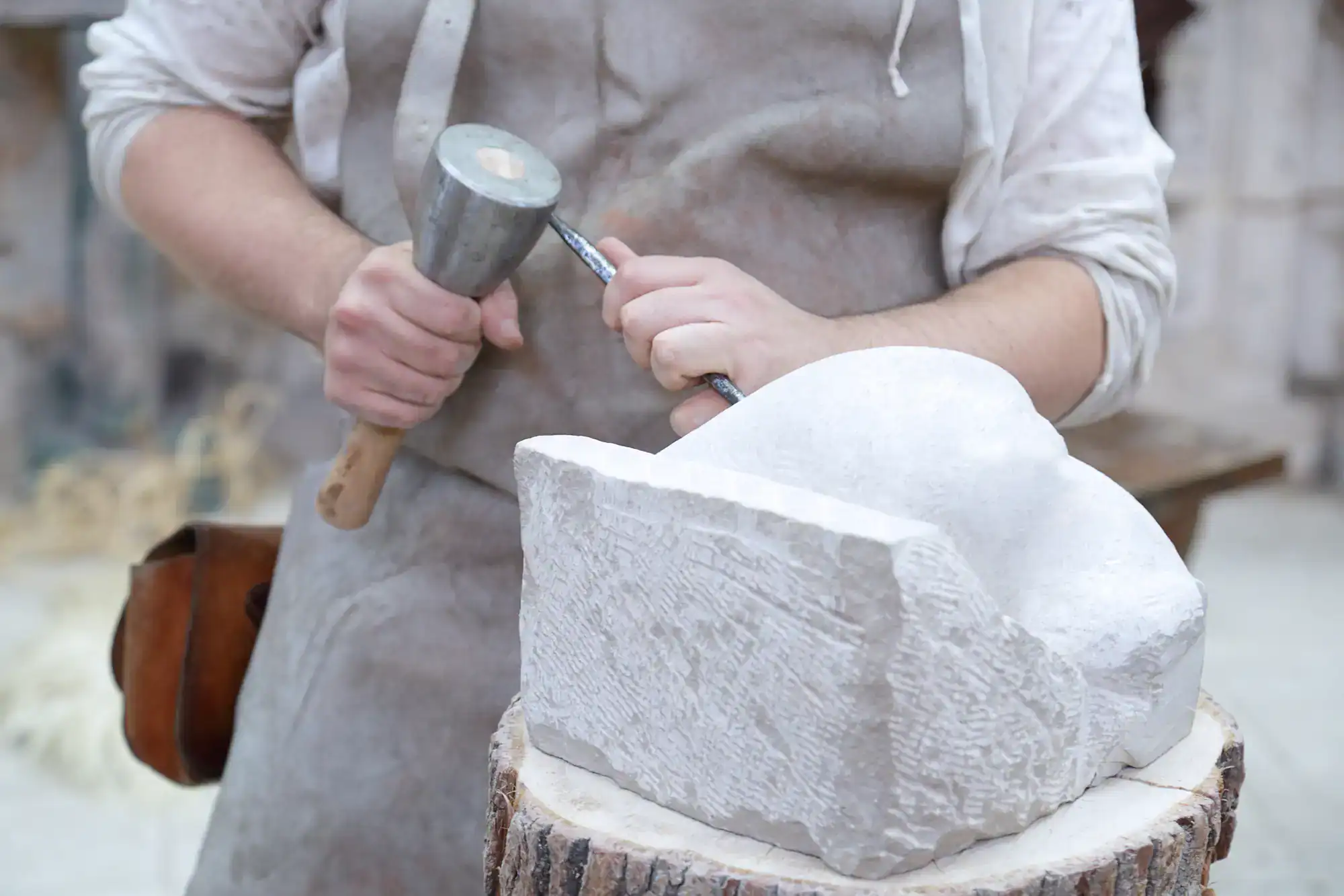 A sculptor wearing an apron uses a hammer and chisel to carve a block of white stone, shaping it into a rounded form. The artist&rsquo;s hands are in focus, with sculpting tools visible on a wooden stump.